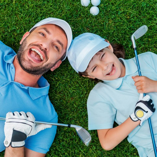 Top view of cheerful little boy and his father holding golf clubs and smiling while lying on the green grass